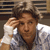 Pixelated static image of a person with brown hair wearing a white shirt and dark tie, hand raised near head against blurred background.