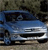 A blue sedan with a silver front bumper and grille is positioned on a textured ground surface under a dark, blurred background.