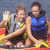 Two people in a red boat on water, one waving with left hand while the other holds yellow oars; background shows blue water.