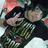A person in a black shirt with green and white patterns makes a peace sign with their right hand, wearing a dark cap, against a light background with orange accents.