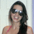 A woman with long dark hair and sunglasses smiles broadly, wearing a red strap top against a light background.