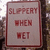 A static pixelated sign with red text reading 'SLIPPERY WHEN WET' on a light gray background, bordered by dark red lines.