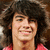 Pixelated close-up of a person with dark curly hair wearing a red garment, smiling broadly against a blurred background.