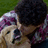A person with dark curly hair wearing a purple and white plaid shirt holds a light brown dog with a black nose and mouth, both facing left against a green background.
