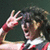 Pixelated image of a person with dark hair making a peace sign gesture, wearing red and white clothing against a dark background.