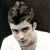 Pixelated static image of a person with dark hair and light skin wearing a white shirt against a dark background, rendered in low-resolution grayscale tones.