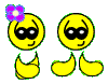 Yellow cartoon character with black eyes and a smile, wearing purple flower headpiece and yellow clothing, shown in two poses against gray background.
