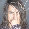 A person with long brown hair and a mustache covers their mouth with their right hand, wearing a dark plaid shirt with red and white stripes against a gray background.