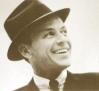 Black-and-white static image of a smiling man in vintage attire with a tilted hat, cropped to show upper body against plain background.