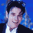 Pixelated static image of a person with dark hair and light skin wearing a white shirt under a black jacket against a blue background.