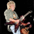 Pixelated image of a person with light hair wearing gray shirt and white pants playing electric guitar against black background.