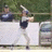 Pixelated static image of a person in athletic attire swinging a bat, with a blurred background of greenery and a figure in the distance.
