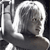 A grayscale pixelated image of a person with short light-colored hair, wearing a dark top, positioned against a dark background with a bright vertical element on the left.
