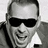 Pixelated grayscale portrait of a person wearing dark sunglasses, a light-colored shirt, and a dark jacket, with a neutral background.