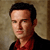Pixelated portrait of a person with short dark hair wearing a red shirt under a dark jacket against a textured beige background.