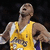 A basketball player in a yellow jersey with purple accents and partially visible text on the chest, arms raised, head tilted back, mouth open, against a dark background.