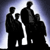 Three silhouetted figures in dark suits stand against a gradient background transitioning from dark blue to white, with one figure holding a briefcase.