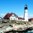 Pixelated static image of a lighthouse with a white tower and black top, red-roofed buildings, rocky terrain, and blue sky background.