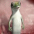 A small lizard with green and white coloring stands on a pinkish surface, surrounded by text reading 'How Can You Drop Him Poor Little guy' in light-colored letters against a dark green background.