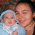 A person holds a baby wrapped in light blue fabric with white trim; the baby wears a matching hooded wrap. The person's hair is dark and short. The background is a blurred brown patterned surface.
