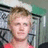 Pixelated static image of a person with light brown hair wearing a red top against a blurred background with green and gray tones.