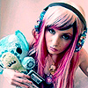A person with long pink hair and blue headphones holds a light blue teddy bear with black eyes and a stitched mouth.