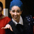 Pixelated static image of a person with blue hair, brown beard, and dark clothing with white collar, holding a white cylindrical object against dark background.