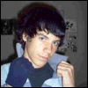 A person with dark hair wearing a dark blue shirt with light blue sleeves sits in front of a wall with small posters, with a heart symbol changing color in the top-right corner.