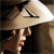 A pixelated figure in profile wears a wide-brimmed straw hat, dark clothing, and a red mouth, with a dark background and light-colored vertical structure.