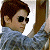 Pixelated image of a person with brown hair wearing dark sunglasses and a light blue shirt holding a brown object.