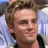 Pixelated close-up of a person with light brown hair, closed eyes, and a slight smile, wearing a dark top against a blurred background.