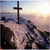 A static image shows a dark cross on a snowy mountain peak at sunrise with the sun low in the sky, next to a red background with yellow text reading "I believe".