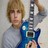 Pixelated image of a person with light brown hair holding a blue and white electric guitar against a neutral background.