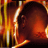 A pixelated figure with dark skin and short hair appears in profile, facing left, against a warm-toned background of orange and yellow vertical stripes.