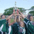 Pixelated image of multiple figures in a group pose with arms raised, set against green foliage and light blue sky background.
