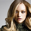 A static close-up of a person with long wavy blonde hair, wearing a dark high-necked top against a plain light background.