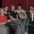 A group of young men in casual clothing sit together against a dark red background with text overlaying the image.