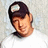 Pixelated image of a person wearing a dark cap with a light circular emblem on the front, set against a light gray background.