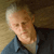 Pixelated image of a person with light skin and short white hair wearing a blue collared shirt against a brown textured background.