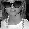 Black-and-white close-up of a person wearing large dark sunglasses, a white top, and a beaded necklace with light-colored spherical beads.