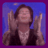 A person in a dark jacket and light shirt performs hand gestures against a blue background with purple border.