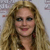 Pixelated portrait of a person with long wavy blonde hair, blue eyes, and a slight smile, wearing a dark top with red accents against a light background.