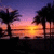 Pixelated sunset scene with orange and yellow sun reflecting on water, dark silhouetted trees on both sides, purple and pink sky gradient.