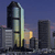 Pixelated cityscape featuring a tall, curved glass building with horizontal bands, surrounded by shorter structures under a dark blue sky with faint pink and purple hues.