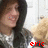 A person with long dark hair wearing a gray jacket over a white shirt holds a light brown teddy bear, with red text and symbols overlaid on the image.