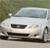 Pixelated image of a white sedan car with dark front grille and headlights, positioned on a light-colored road against a blurred green background.