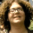 A pixelated close-up of a person with long dark hair, wearing glasses, and smiling, set against a blurred background with warm tones.