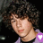 Pixelated close-up of a person with curly dark hair, light skin, and a purple garment, featuring a small purple and white symbol on the right shoulder.
