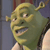 Pixelated green ogre face with large eyes, small nose, and open mouth showing teeth, set against a blurred background with blue and brown tones.