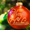 A red spherical Christmas ornament with 'Merry Christmas' text in gold script, hanging from a tree with blurred green and yellow lights in the background.